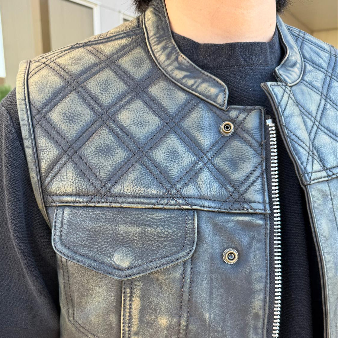 A close-up of a person wearing a distressed black leather vest with diamond-quilted shoulder panels, a stand collar, a front zipper, and buttoned chest pockets over a black t-shirt.