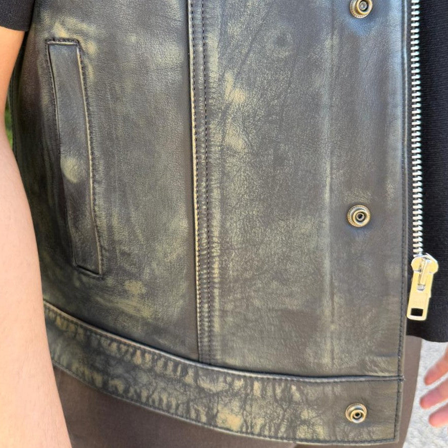 A close-up of a person wearing a distressed black leather vest over a black shirt, showing the front zipper, snap buttons, and side pocket.