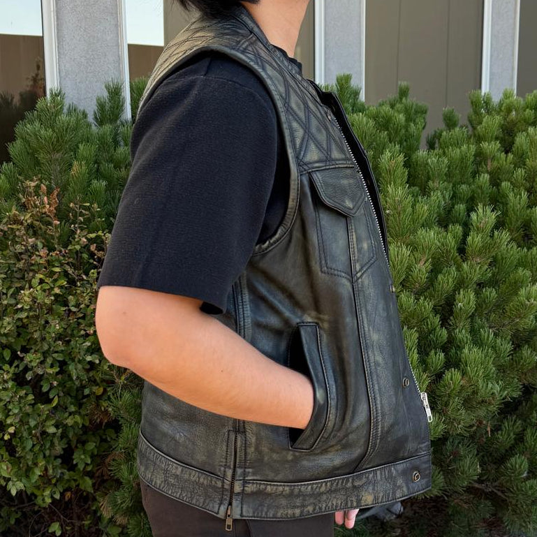 A person standing outdoors in profile wearing a dark distressed leather vest over a black t-shirt and dark pants, with green bushes and a building in the background.