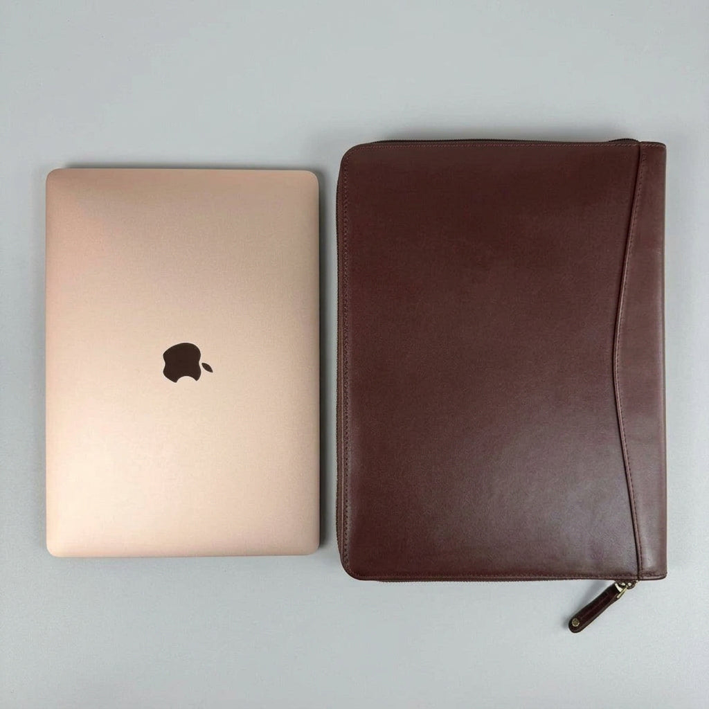 A brown leather zippered laptop sleeve shown next to a closed gold Apple MacBook