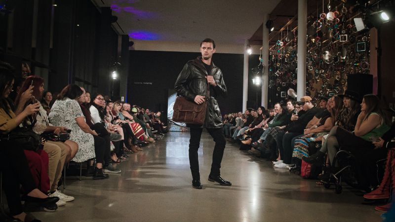 A male model walks a brightly lit runway at a fashion show, wearing a mid-length black leather jacket. He carries a large, dark brown leather messenger bag over his shoulder.