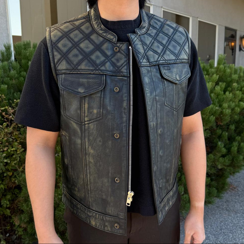 A man wearing a distressed black leather motorcycle vest over a black t-shirt and dark pants, standing outdoors on a paved walkway in front of a building.