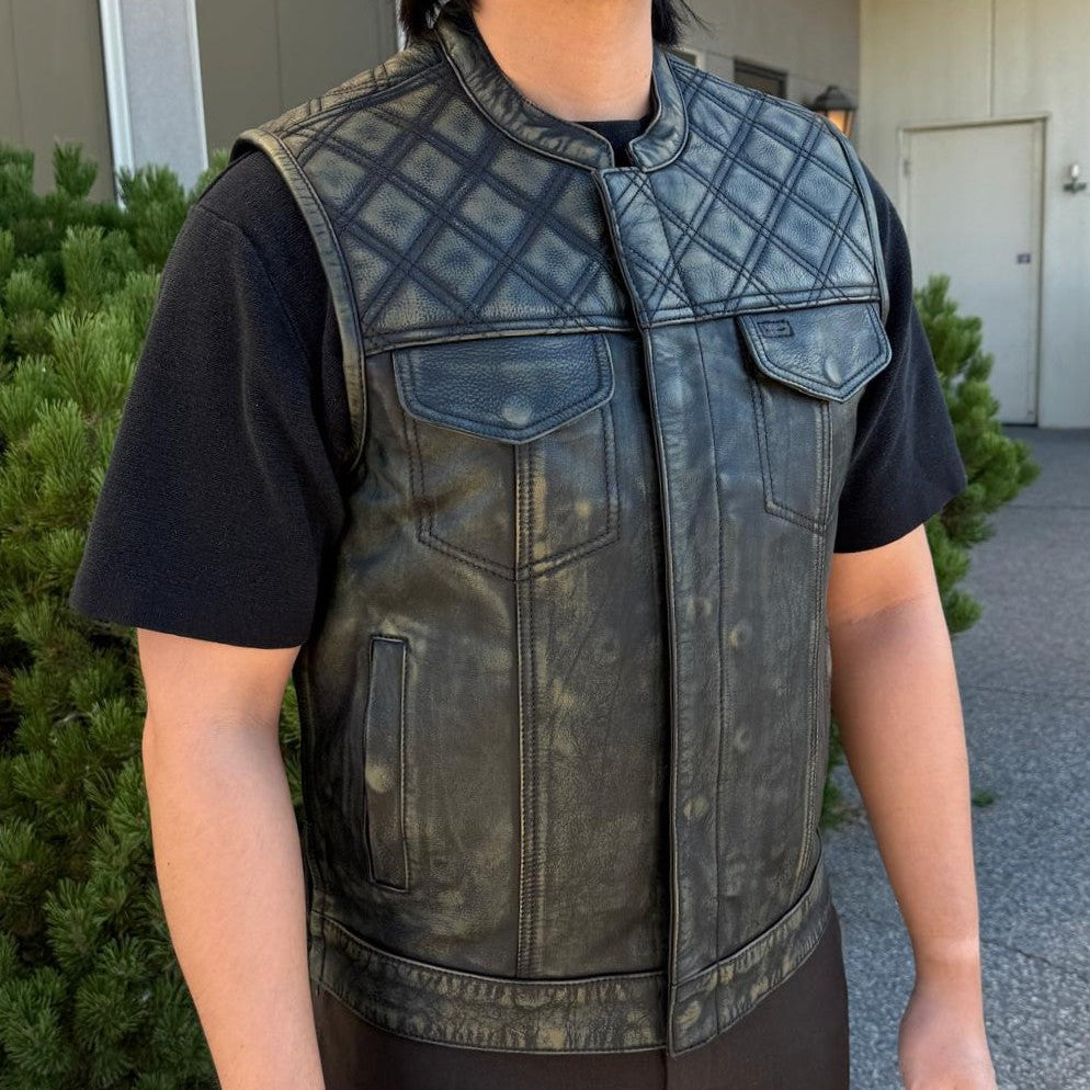 A man wearing a distressed black leather motorcycle vest over a black t-shirt and dark pants, standing outdoors in front of a building.