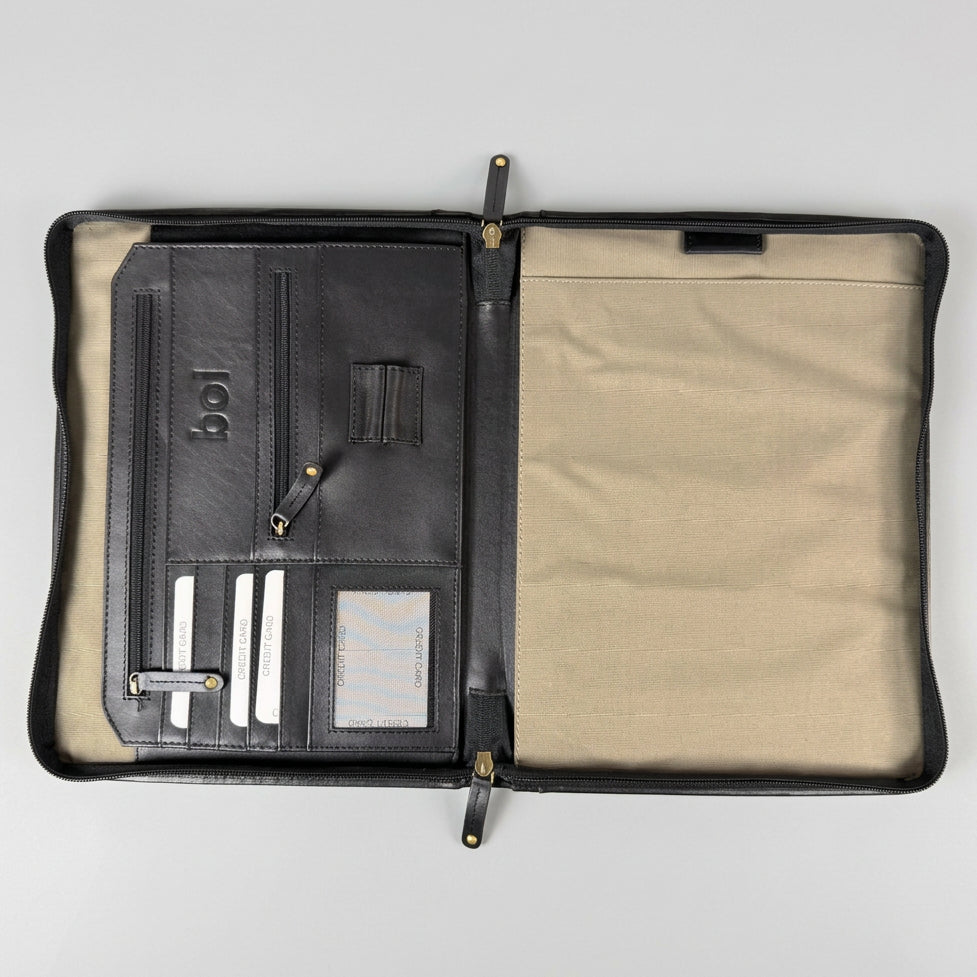An open black leather organizer case with a beige interior, laid flat on a gray wood-textured surface. The left side shows card slots, a zip pocket, elastic loops, and a small hanging tag, while the right side is a plain beige fabric panel.